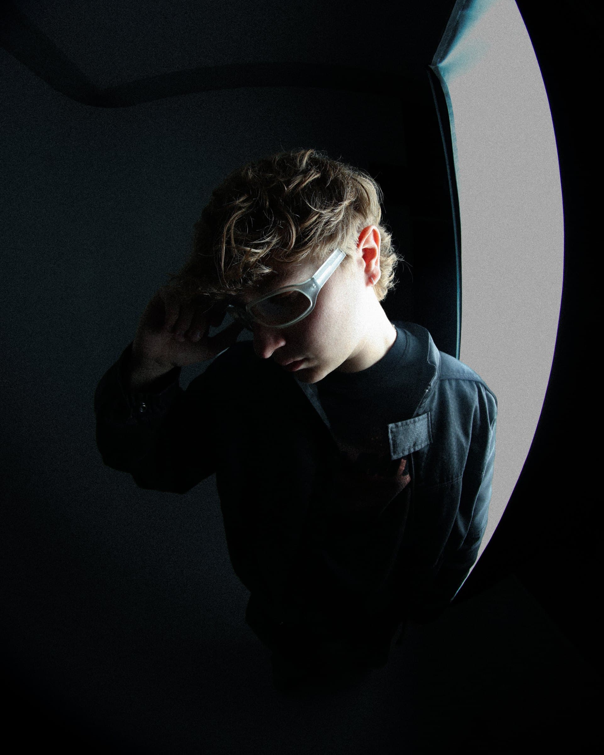 Bruno Brero in clear sunglasses, looking down, lit from the side in a dark room